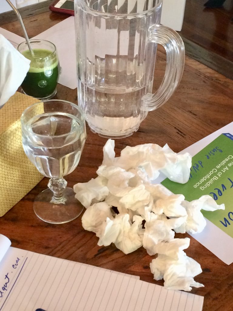 Table top covered with a full water glass, half-full water pitcher, cup of green juice, crumpled tissues and a notepad.
