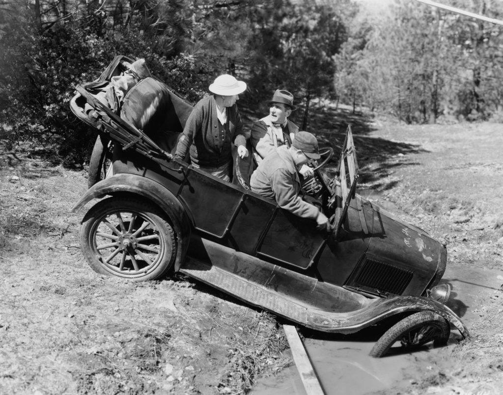 Black and white image of an old-fashion vehicle stuck in the mud with 3 passengers.