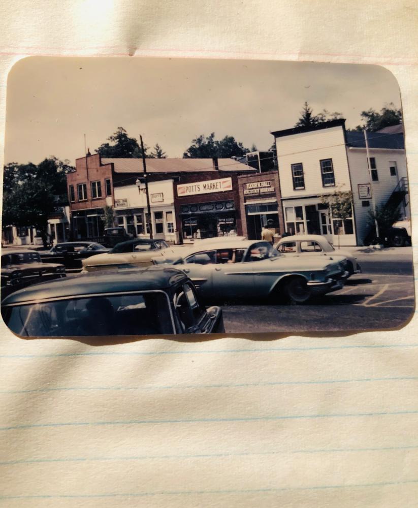 An old photograph of a town street lined with cars.