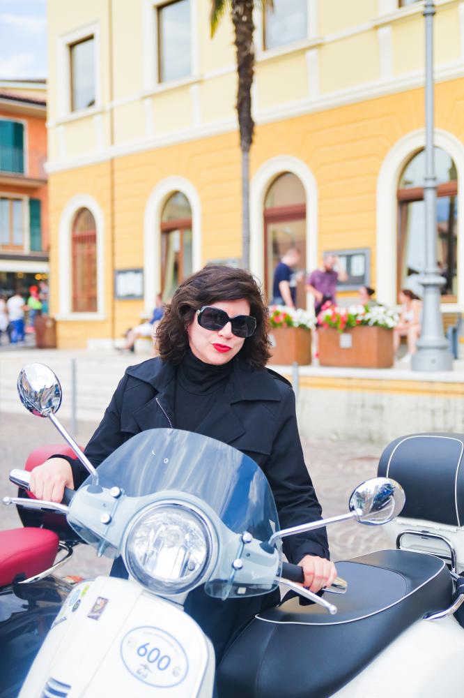 Susie deVille wearing a black jacket, black sunglasses and red lipstick sits on a moped. 