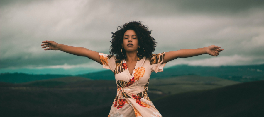 Female presenting person with afro and colorful dress is standing on a mountain with their eyes closed and arms spread, as though feeling it out