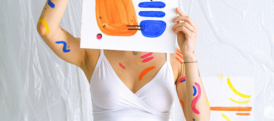 Cropped photo of a person holding a colorful abstract painting in front of their face, with paint covering their arms and chest, to represent how to not be afraid of rejection through creative rebellion