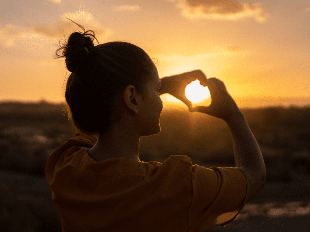 Photo of person silhouetted against golden sunset with their hands held in a heart shape, representing ideas of self love and having your own back 