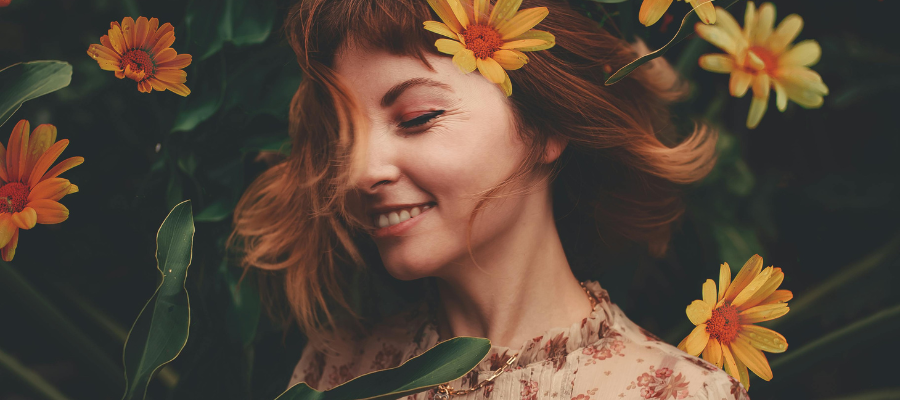 Close-up of a person with their eyes closed smiling and twirling in front of a bush of bright yellow flowers, illustrating mindfulness for positive self change.