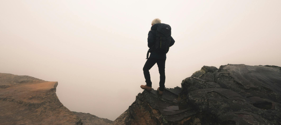 Photo of person wearing a backpack and standing on cliff edge looking out into fog—representing uncertainty and the importance of learning how to handle risk