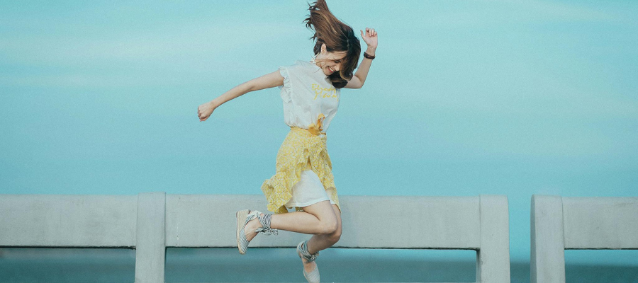 Photo of a person wearing a dress and heels jumping up on a dock with their hair flying and a smile on their face—representing the act of doing something just for fun to overcome self-doubt.