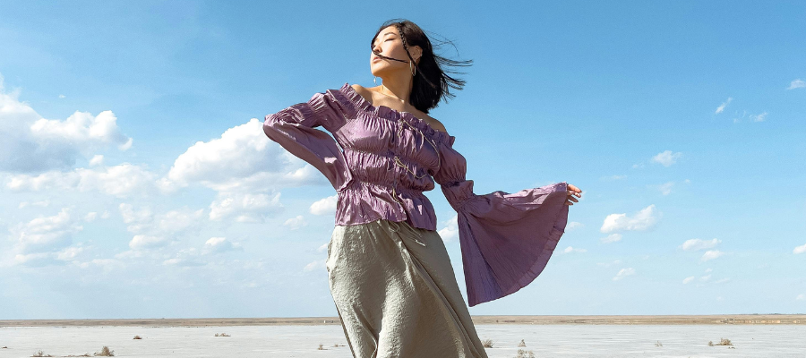 Photo of a person wearing a flowing purple blouse, standing tall, and facing into the wind with their hair blowing back—representing embracing challenges, taking ownership, and achieving self-empowerment.