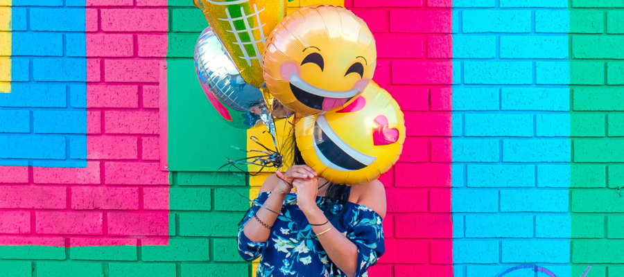 A woman holds colorful balloons, including emojis, in front of her face while standing in front of colorfully painted brick to symbolize a creative, effective entrepreneurial mindset.