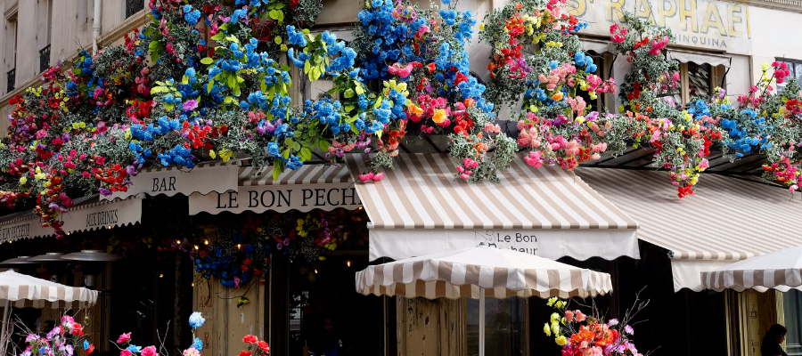 Exterior of Le Bon Pêcheur, a cafe in Paris, covered in a colorful array of flowers. Blue, pink, green and red flowers cover a striped awning over the entrance. Representing French brands