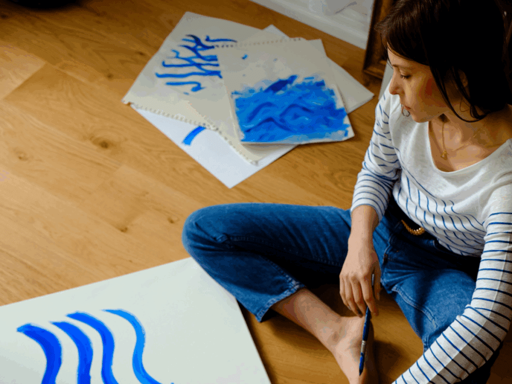 Woman sits on the floor with a work in progress painting of blue lines, work discarded to the side. Her face is downcast, as if she is filled with self doubt.