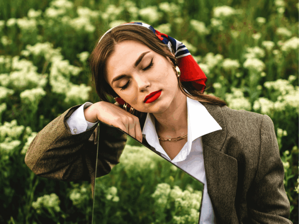 person with eyes closed in a field with bright red lipstick and a red and blue scarf in their hair, wearing a business jacket and leaning against a mirror with a blissful smile on their face, to symbolize how to be yourself