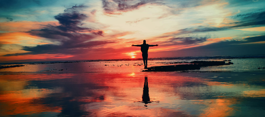 letting go; a person standing on a sandbar during low tide with arms outstretched in front of a beautiful sunset to symbolize letting go.
