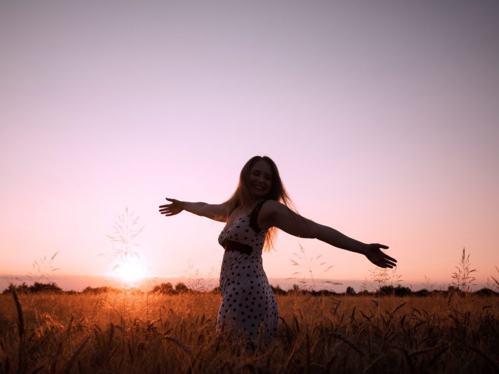 A woman stands in an open field, the sun setting behind her. Her arms are outstretched and she smiles, representing giving herself permission to want, letting go of self disappointment