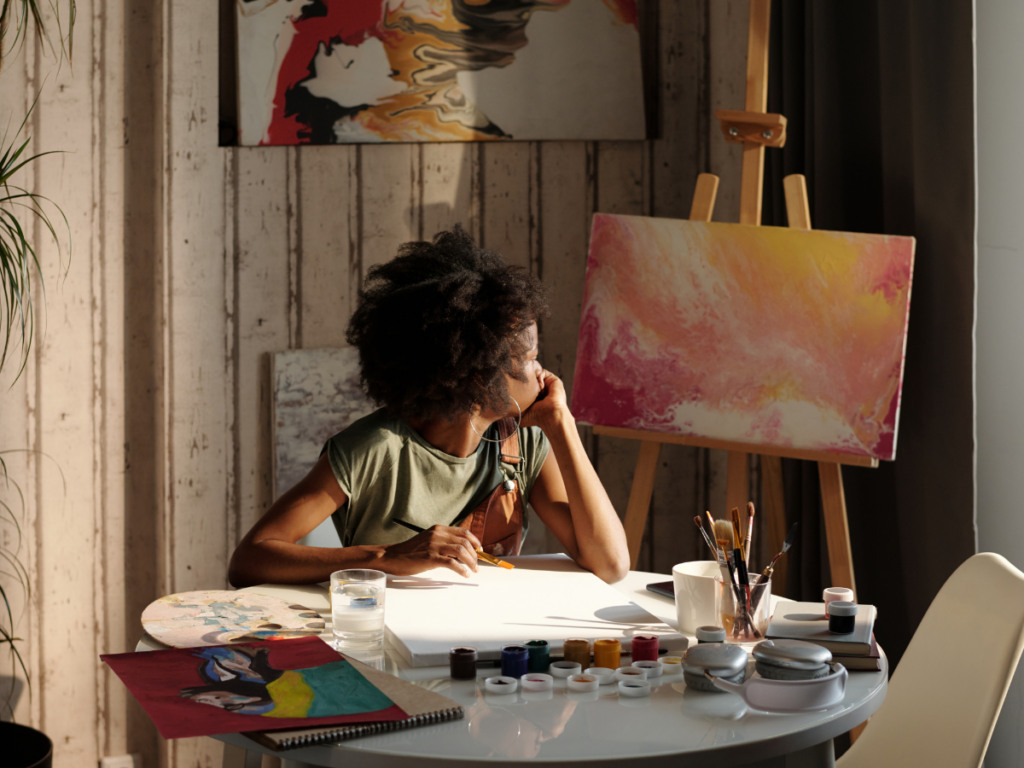 A woman sits at a blank canvas, looking over her shoulder at her previous work, a pink and yellow abstract painting, hesitating to begin her next work, representing the fear of self disappointment
