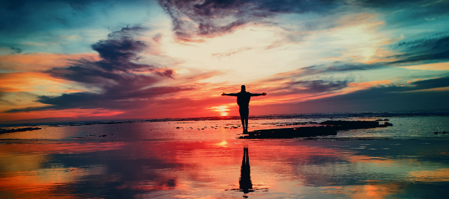 Person stands along a colorful shoreline with a sunset in the background, arms outstretched, representing letting go of disappointment