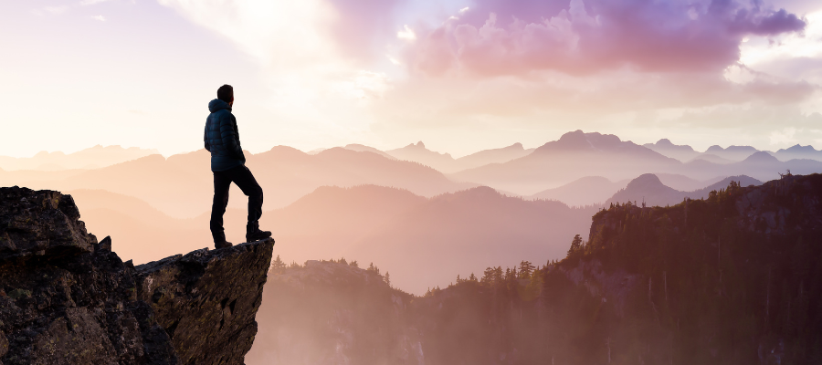 Person standing on cliff with mountains and sunset in background. Standing on the edge of the unknown.