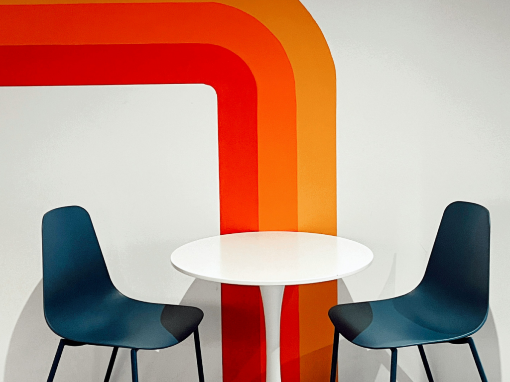 A simple table with two empty chairs sits against a wall painted with bold red, orange, and yellow stripes that lead directly down to the seating area, creating a space to invite more creativity.