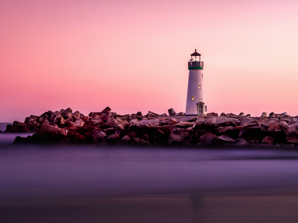 photo of a lighthouse on a rocky outcropping against a pink and purple sunset represents what creators and entrepreneurs needs to be for their audiences: a lighthouse, a guide to make them feel seen. 