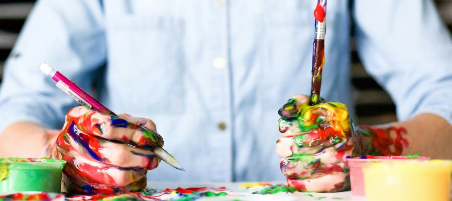 Photo of a person seated at a desk with their hands covered in paint holding a paintbrush and a pencil represents how messy, pressure-free play can help us unlock our imagination, trust our intuition, and get past creative block.
