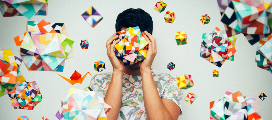 An artist holding a colorful geodesic paper ball they created while similar balls float around them represents finding serenity and calm focus when faced with the pressure to do more.