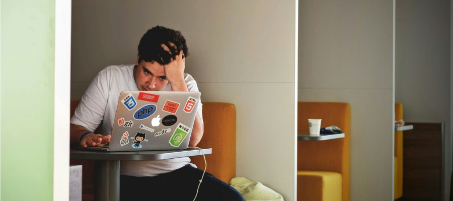 photo of a man with a hand in his hair looking frustratedly at a laptop as he sits in a cafe booth represents the hidden friction that keeps us from great work